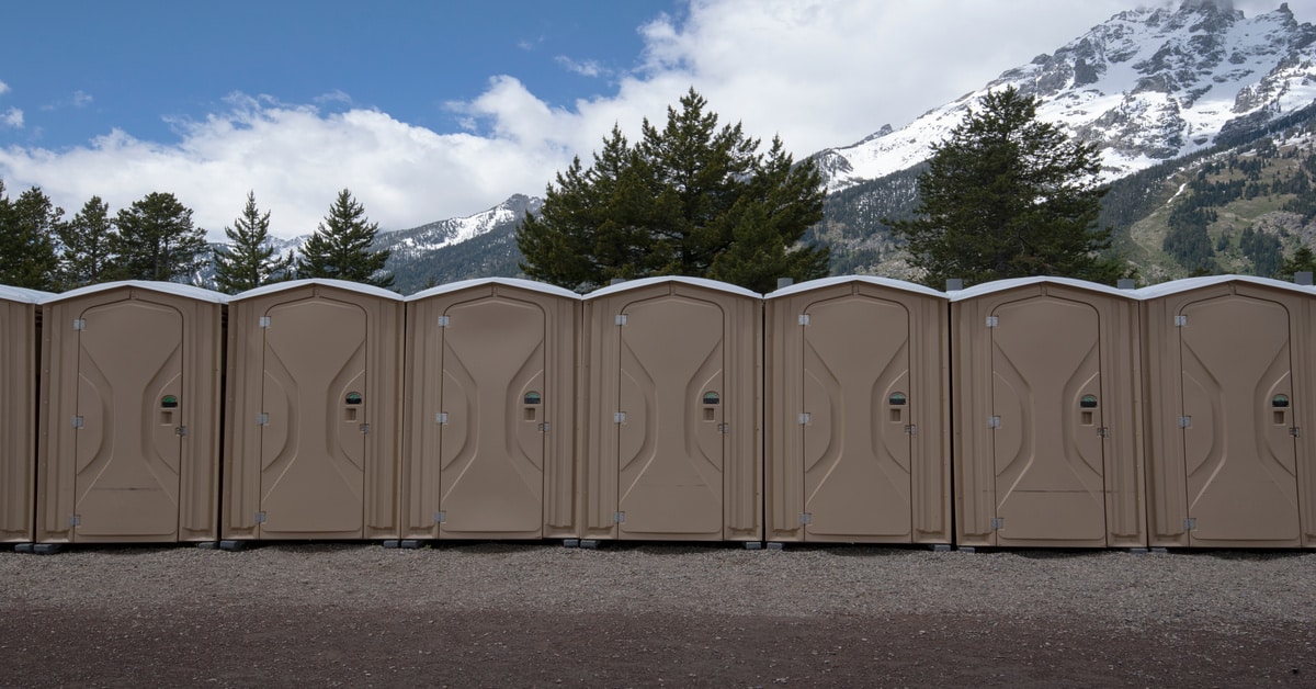 A line of portable toilet stalls with fully enclosed spaces for people. These units are on a gravel road.