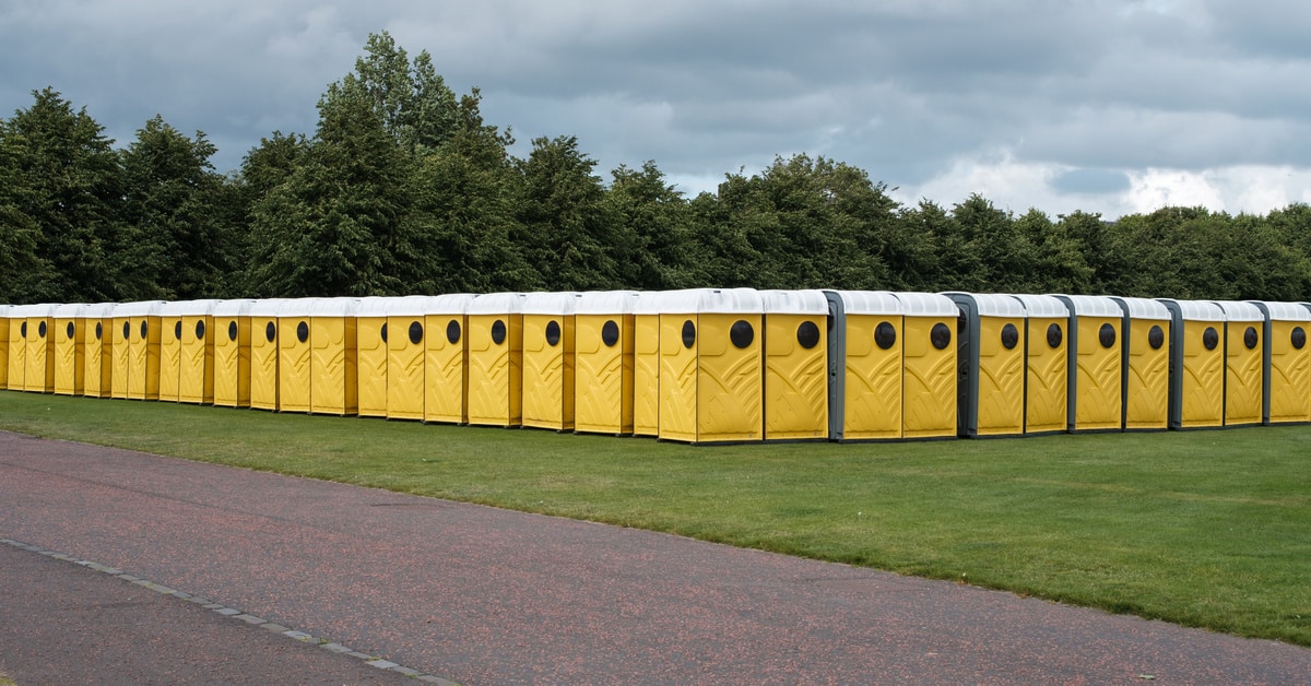 Two rows of portable toilets that meet at the corner of each line. The stalls are sitting on grass next to the road.