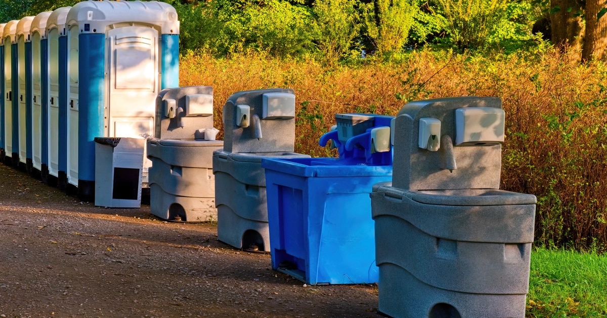 Portable hand washing stations and restrooms are at an outdoor event site with trees in the background.