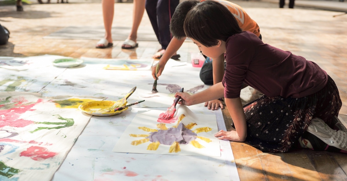 A group of children paint colorful artwork on a large sheet of paper at an outdoor event with messy hands and paint supplies.