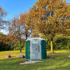 A series of portable bathrooms positioned in the middle of a grassy field. There is a single tree right behind the bathrooms.
