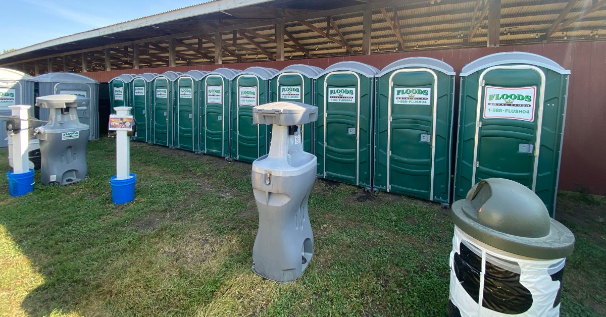A line of porta potties set up outside at an event. There are also a couple of portable hand washing stations.