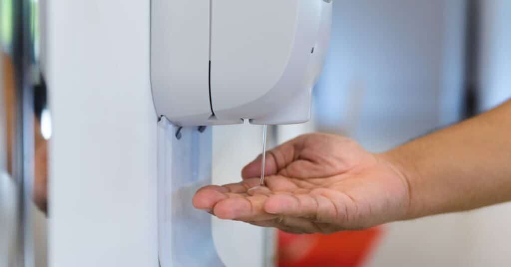A close-up view shows a hand underneath an automatic hand sanitizer pump. Sanitizer is flowing from the pump.