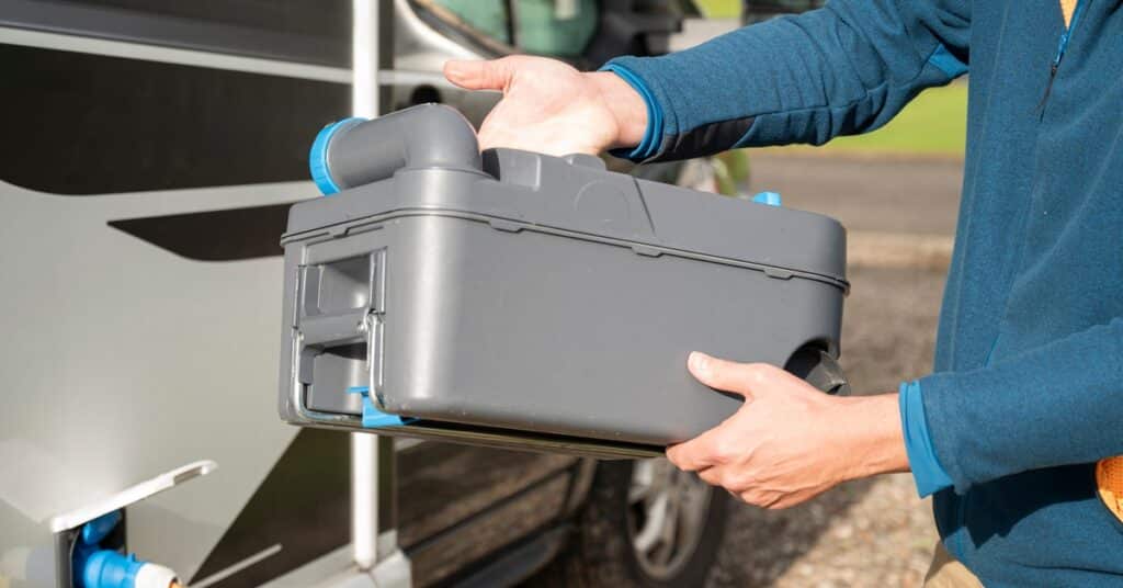 A person wearing a gray and teal lightweight jacket holds a holding tank next to a gray and black RV.