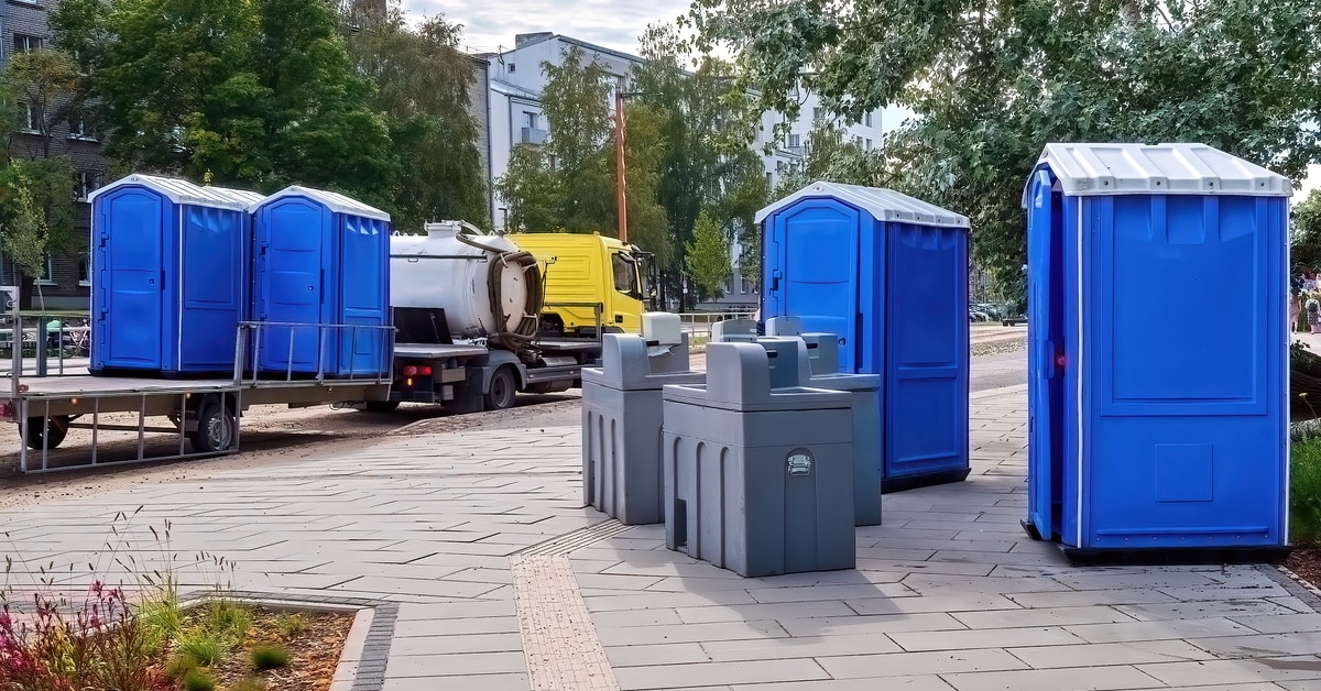 Two blue porta-potties sit on a sidewalk next to three gray portable holding tanks. A truck appears on the road.