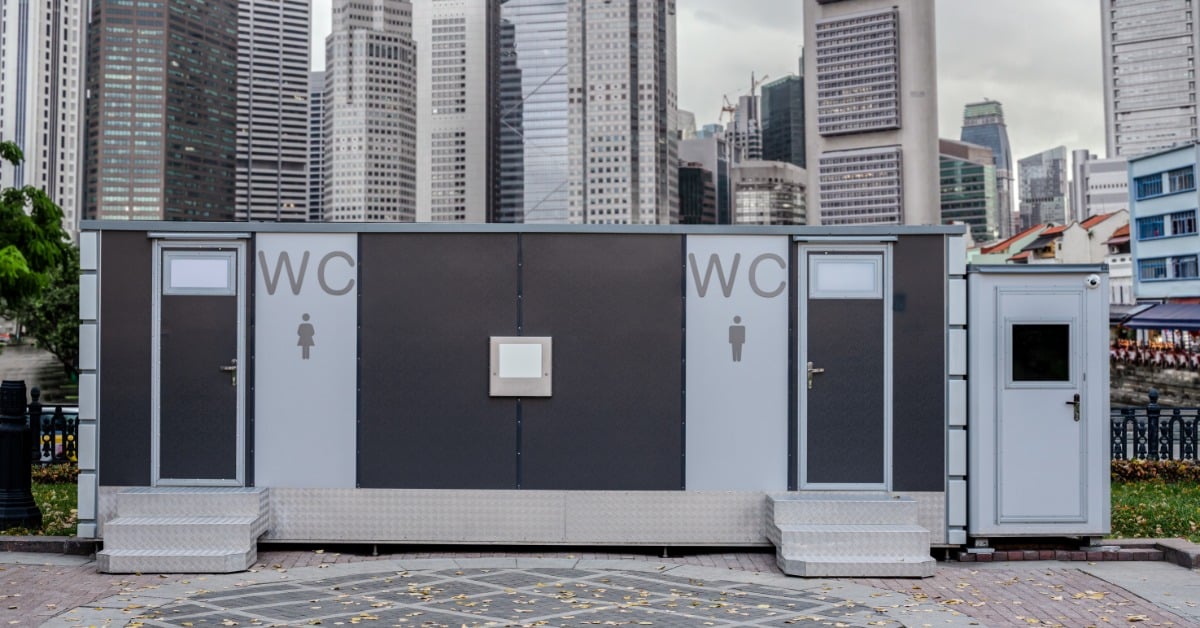 A modern outdoor restroom with steps leading up to the doors. There is a large city behind the trailer.