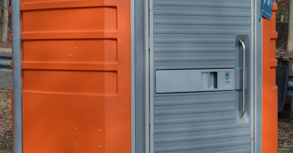 A large ADA-compliant porta-potty with orange, white, and grey accents is outside in a public space.