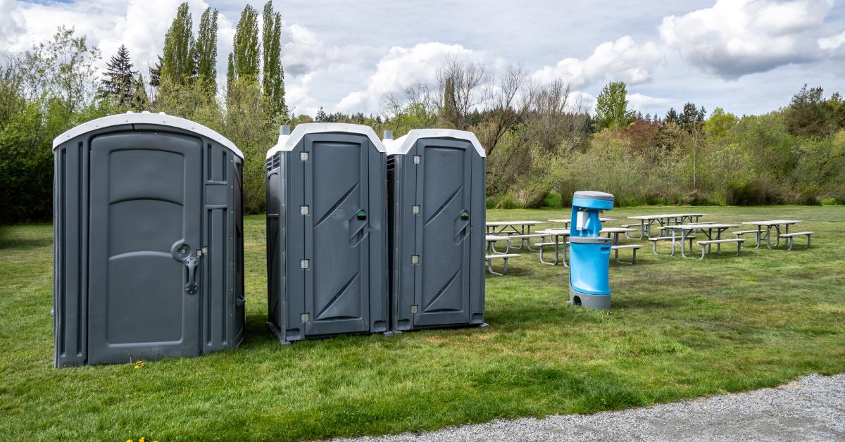 A light blue handwashing station sits next to three portable restroom units on the grass at an outdoor park.