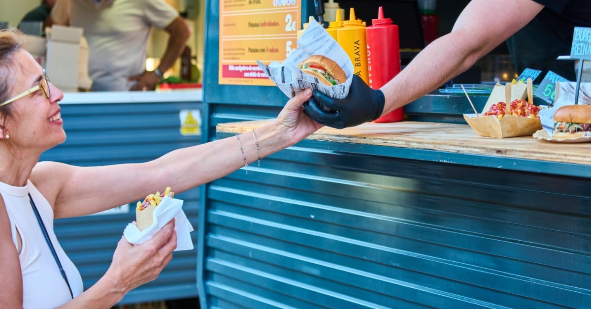 A vendor wearing gloves hands a sandwich to a customer at an outdoor food market booth with condiments and a menu board visible.