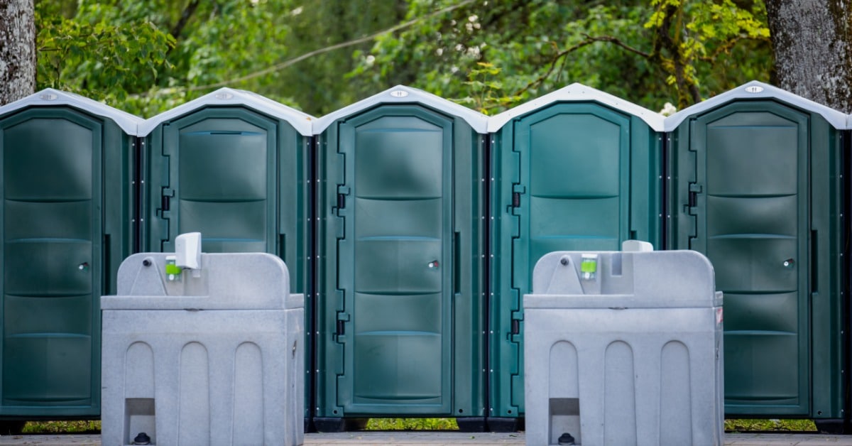 A row of green portable restroom units with two gray handwashing stations set up outdoors near trees at an event site.