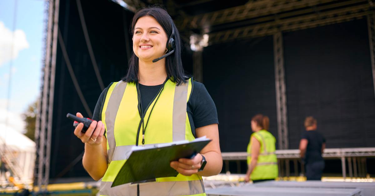 An outdoor concert coordinator in a high-visibility vest holds a clipboard and radio while managing event logistics near the stage.