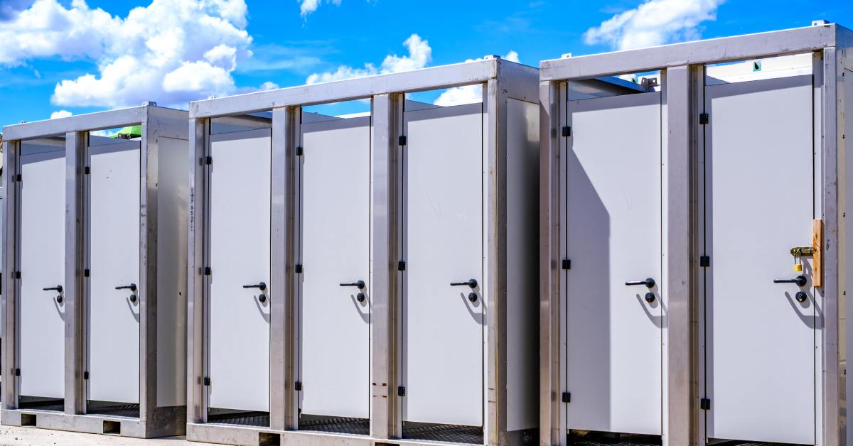 A row of portable restroom units lined up outdoors under a bright blue sky, set up for an event or large gathering.