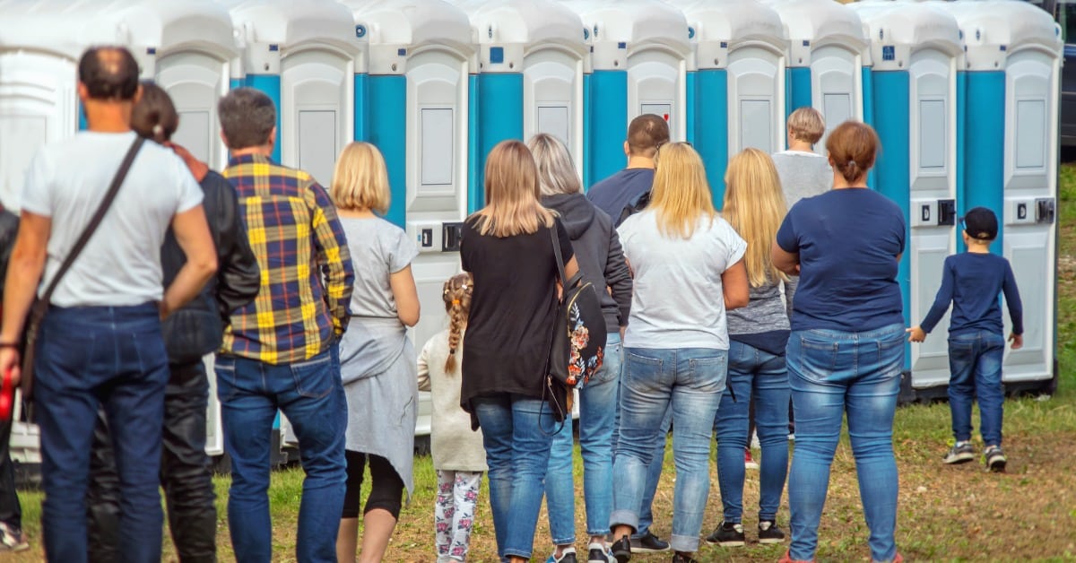 A crowd of adults and children seen from behind, waiting in line for a row of white and blue portable toilets outdoors.
