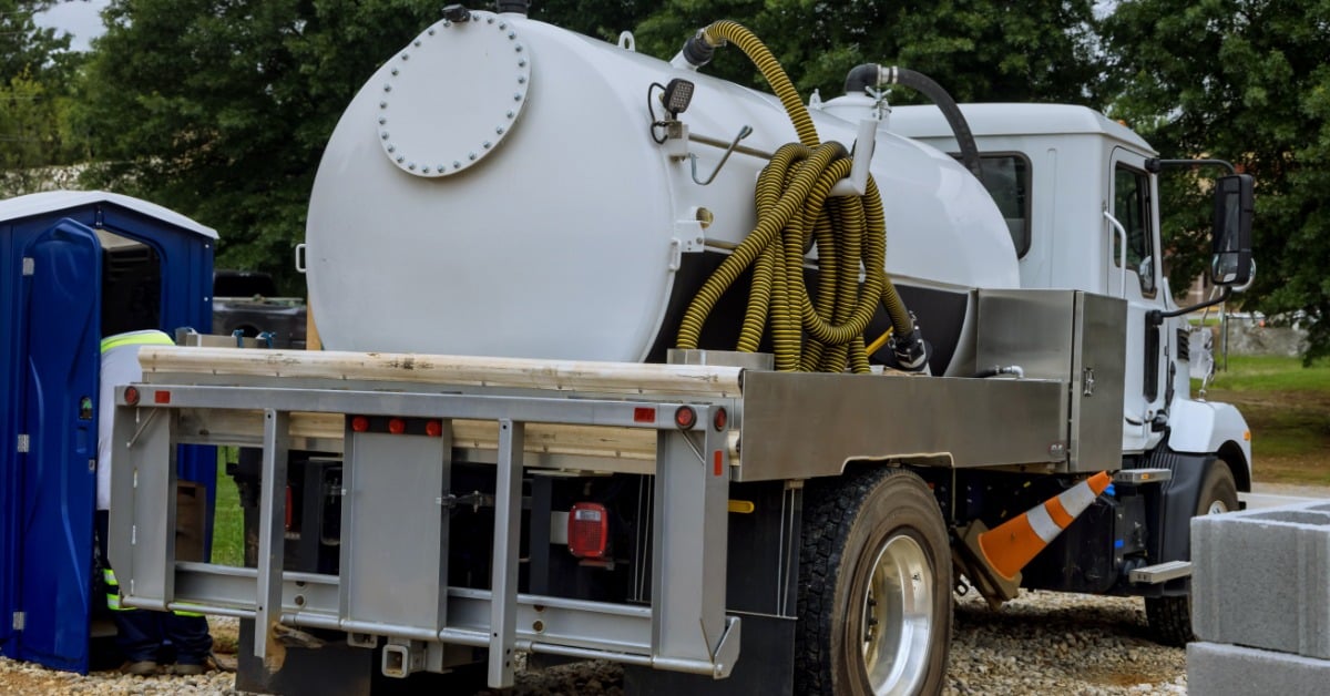A white septic service truck with yellow coiled hoses on a gravel lot, with a blue portable toilet visible in the background.