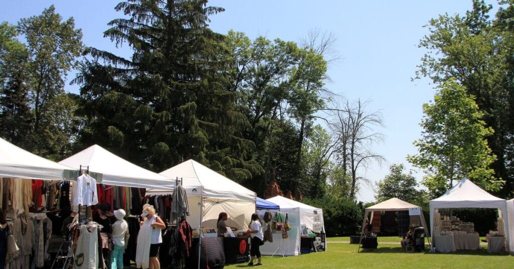 White vendor tents with clothing racks and other items are set up on a grassy park lawn during an outdoor market event.