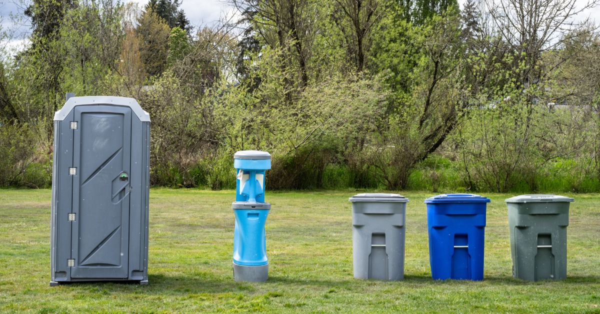 An outdoor rental setup with a portable restroom, a portable hand washing station, and recycling bins on the grass.
