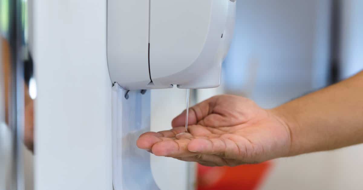 A close-up view shows a hand underneath an automatic hand sanitizer pump. Sanitizer is flowing from the pump.