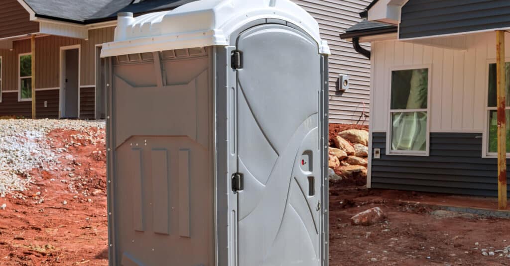 A gray porta potty on red dirt at a construction site. Partially constructed homes are in the background.