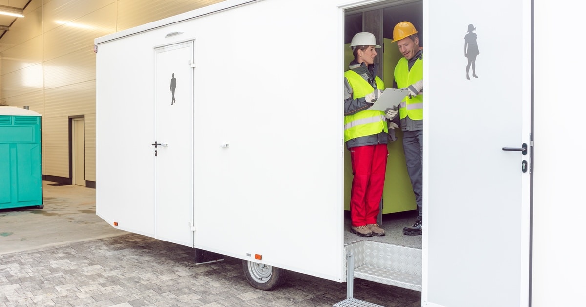 Two workers in construction vests, hard hats, and gloves inspect the inside of a large bathroom trailer.