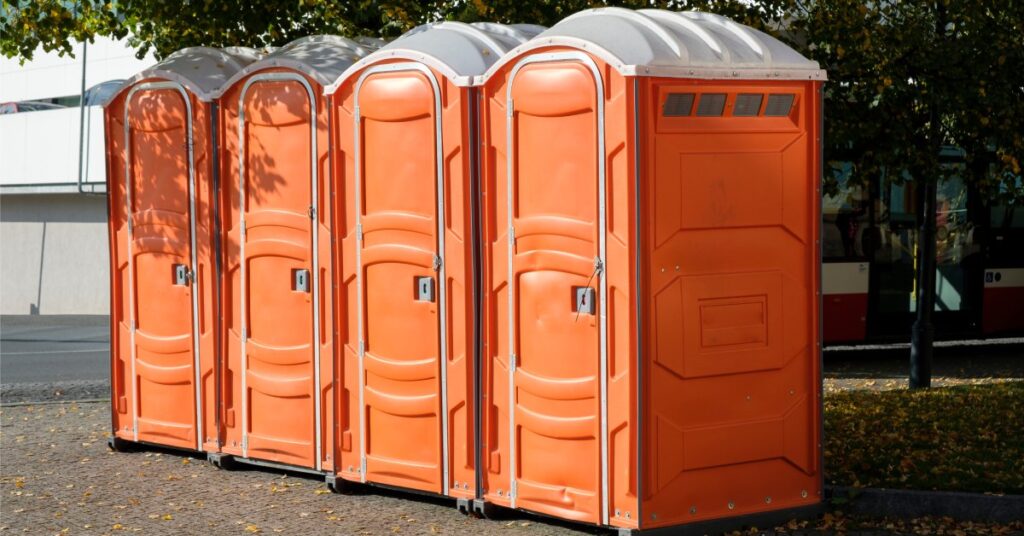 Four orange porta potties sit in a row outside on asphalt. The toilets are positioned under trees, which provide shade.