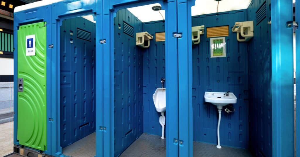 Multiple connected portable toilets sit outside. Three are open to view, and one has a closed green door.