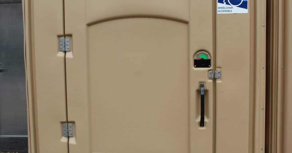 A tan porta potty with silver hinges and a large blue-and-white sticker reading "wheelchair accessible."
