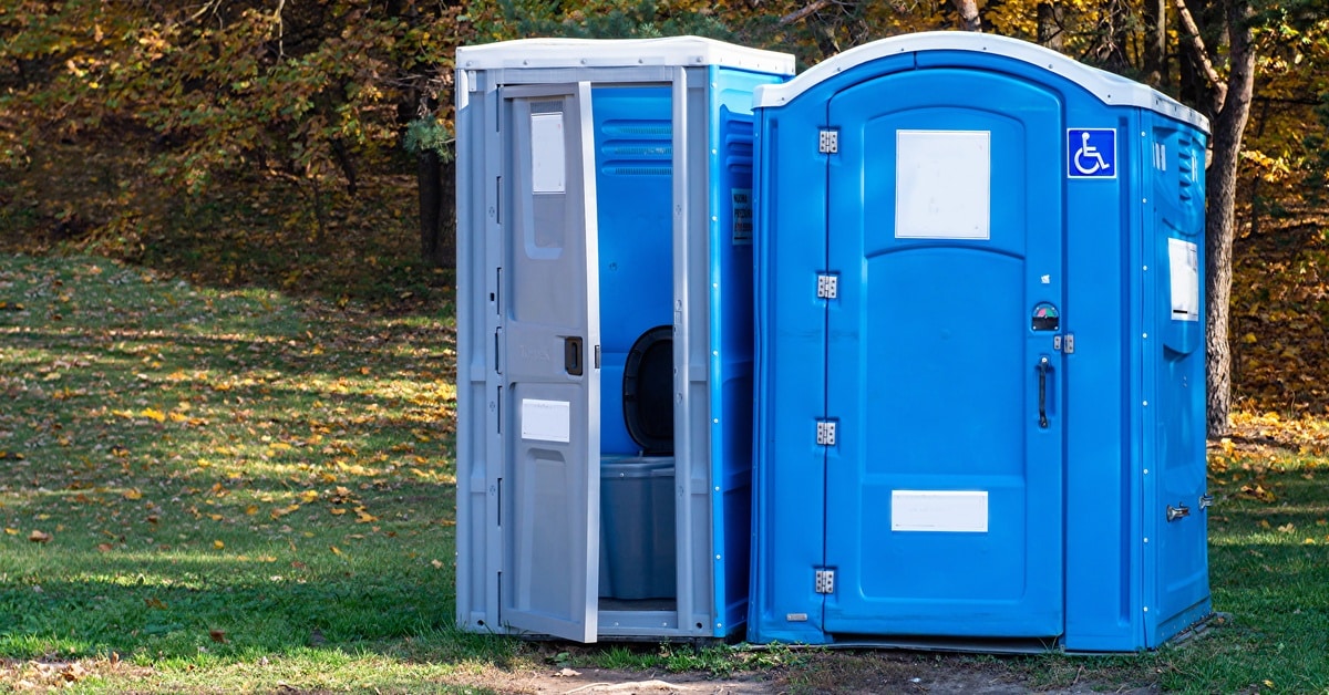 Two porta potties in a park with trees in the background. One unit is slightly larger and has a handicap sticker.