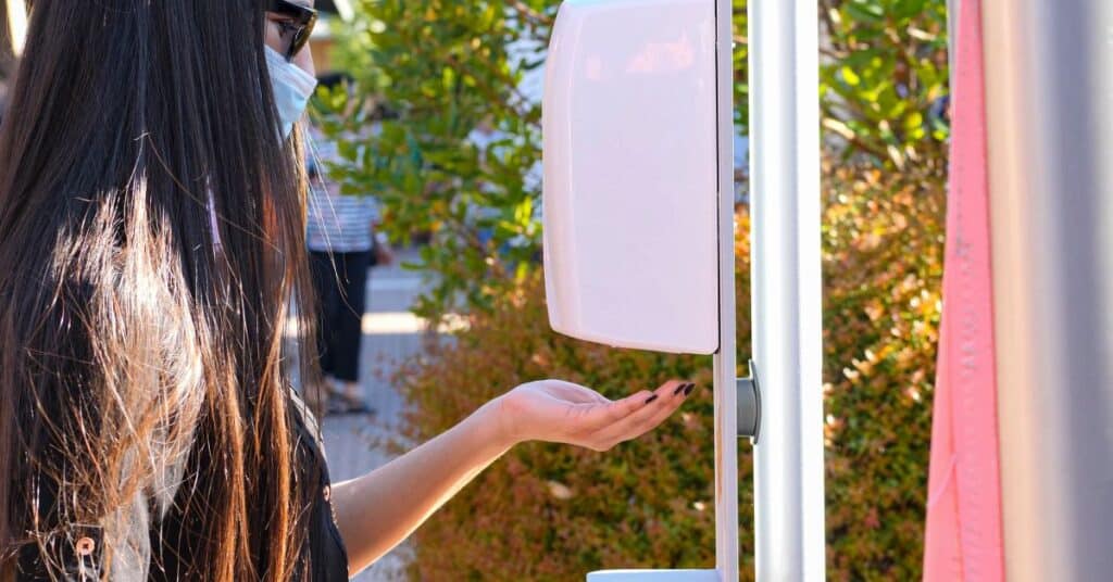 A dark-haired young woman in sunglasses and a face mask getting hand sanitizer at an outdoor dispenser.