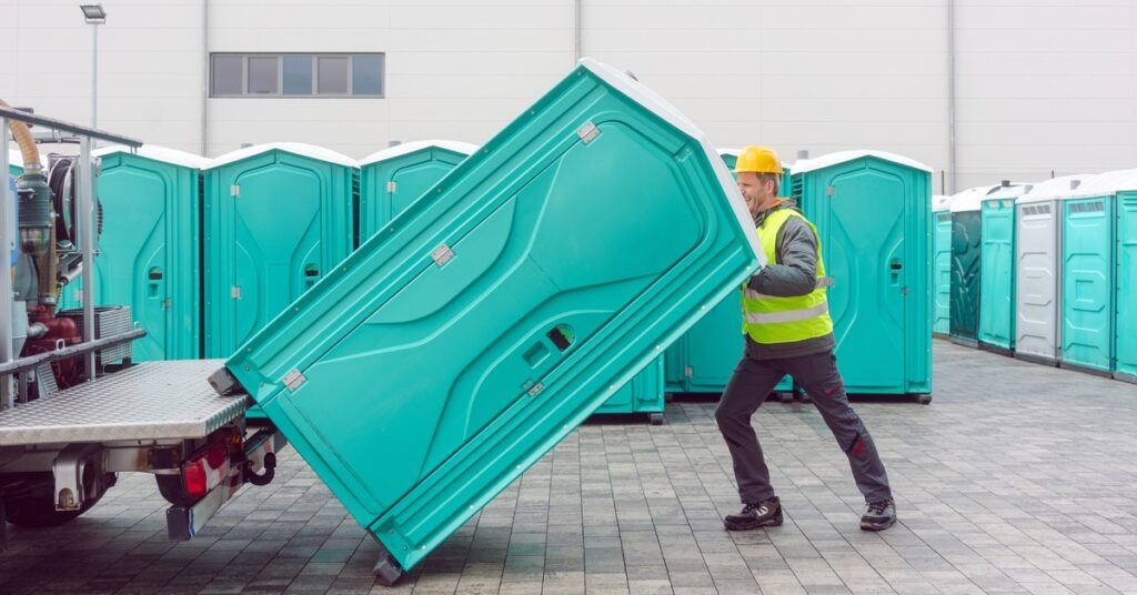 A man in a yellow safety vest loading a porta potty onto a truck. He's standing near rows of porta potties.