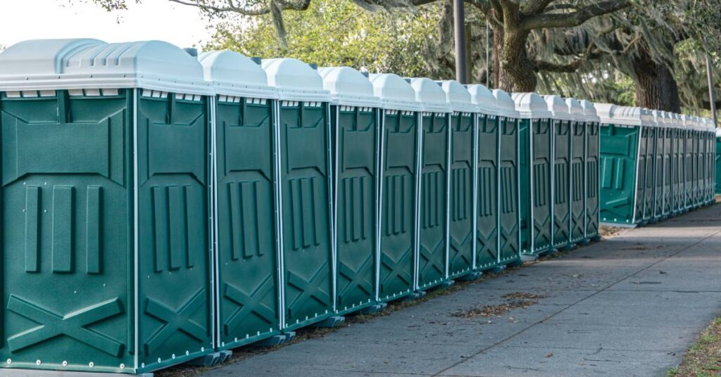 A long row of green portable toilets placed side by side next to an empty sidewalk and towering trees.