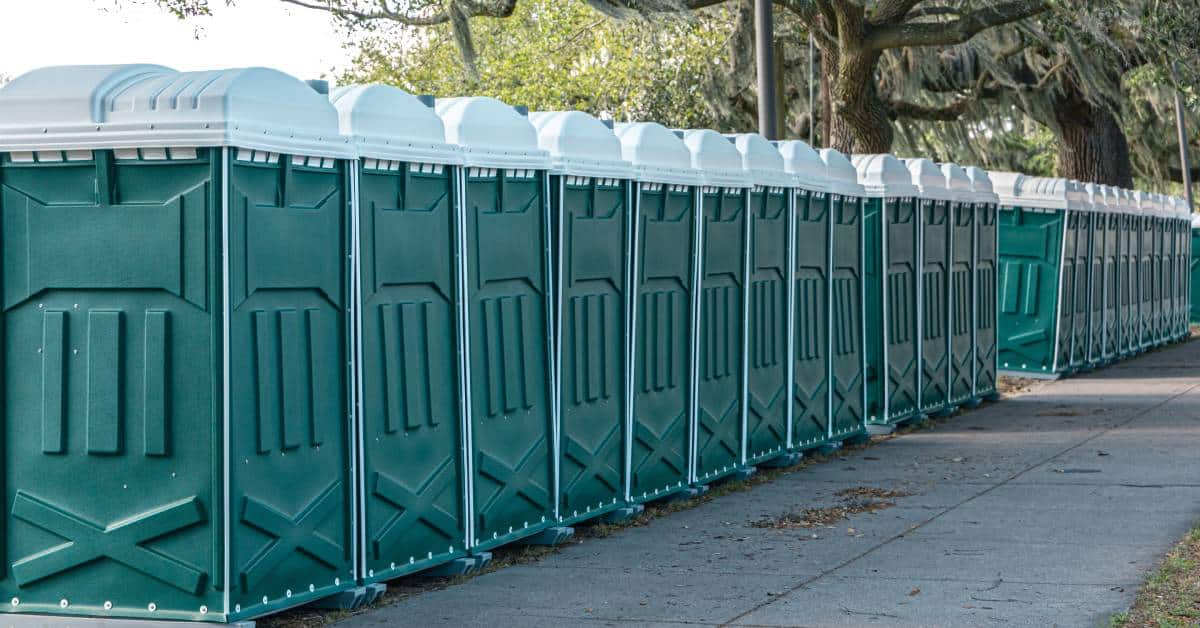 A long row of green portable toilets placed side by side next to an empty sidewalk and towering trees.