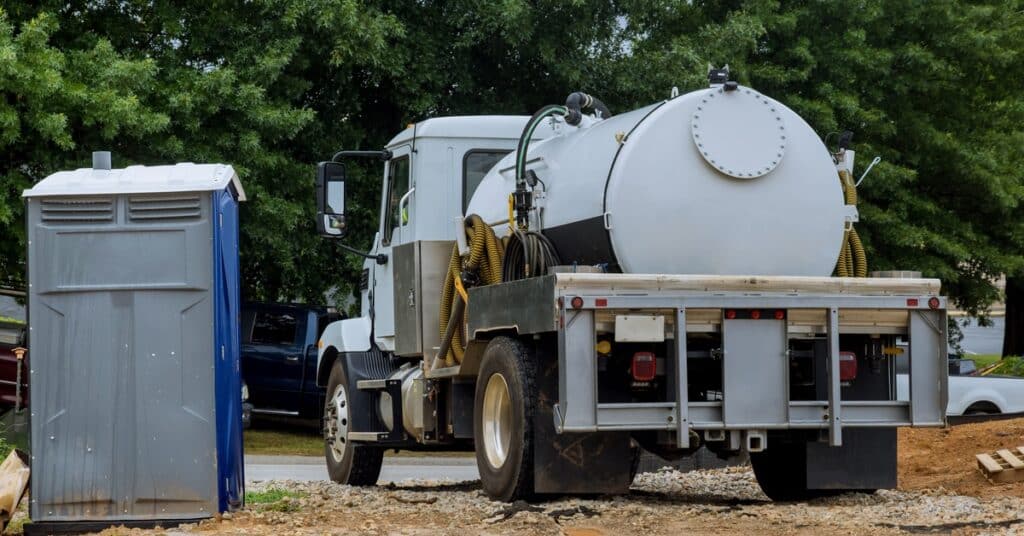 A white septic vacuum truck parked in a dirt field next to a small portable toilet and multiple trees.