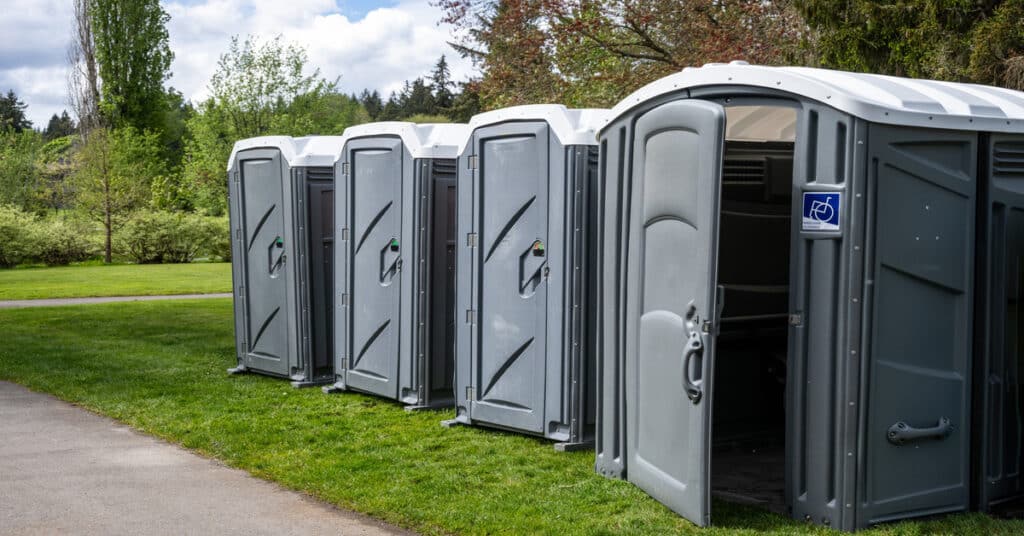Four gray-and-white portable toilets arranged on a grassy area. One toilet has an ADA sticker and an open door.