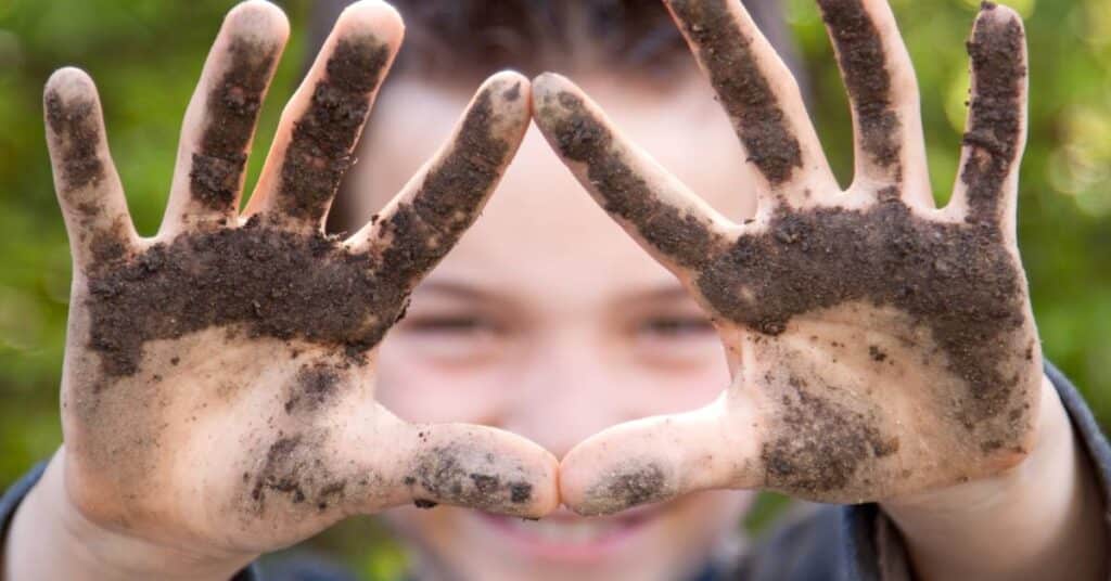 A grinning young boy holding out his hands, which are covered in fresh dirt. Blurred greenery is visible behind him.