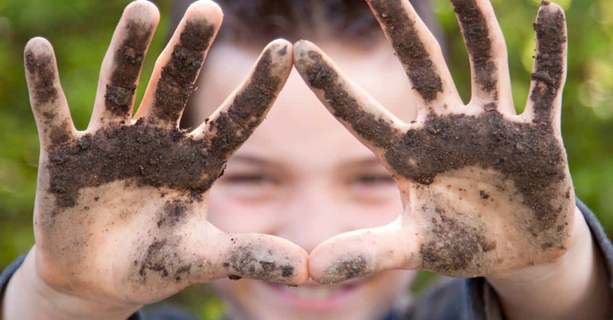 A grinning young boy holding out his hands, which are covered in fresh dirt. Blurred greenery is visible behind him.