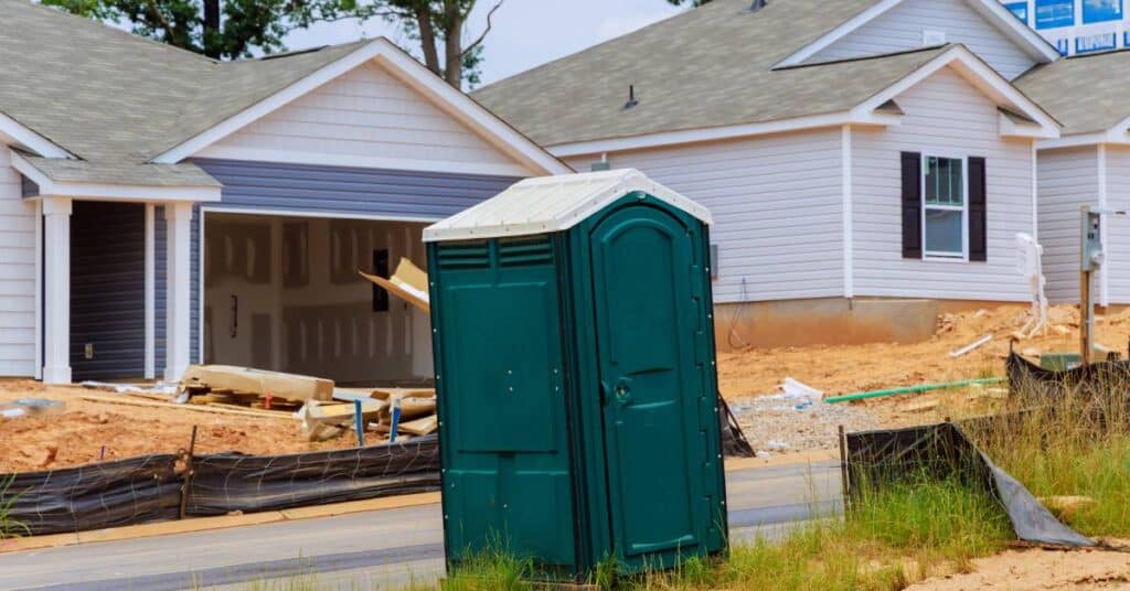 A green portable toilet with a white roof positioned by the side of the road at a residential construction site.