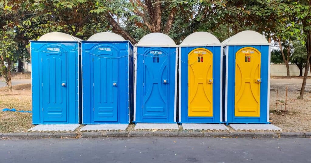Five blue portable toilets positioned in a row by the side of a road. Two toilets have yellow doors.