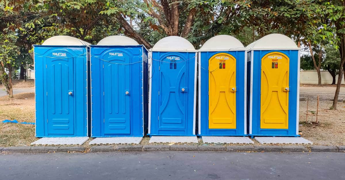 Five blue portable toilets positioned in a row by the side of a road. Two toilets have yellow doors.