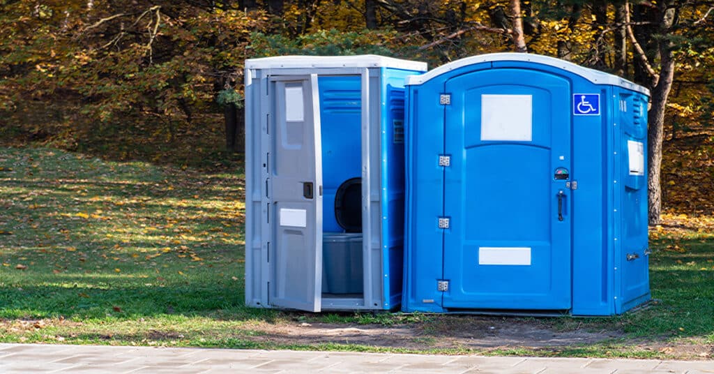 Two blue portable toilets positioned side-by-side in a grassy area. One of the toilets has an ADA sticker.