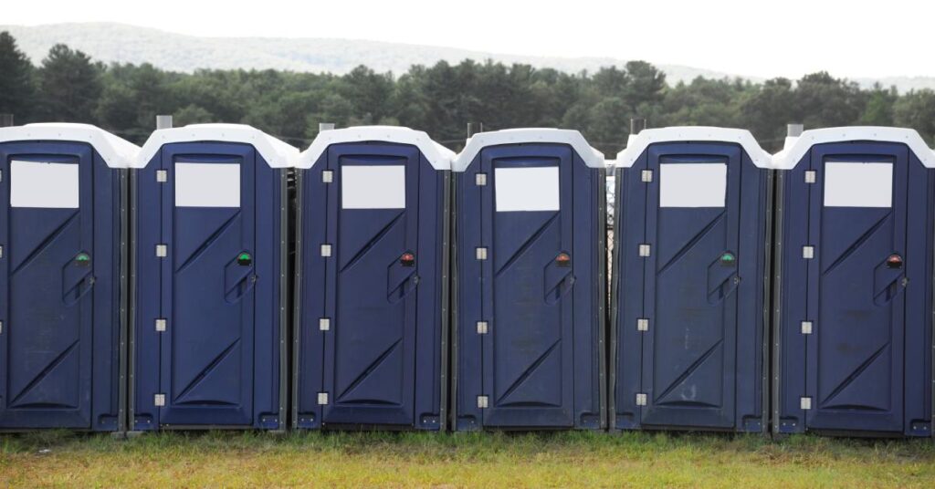 A row of six navy-blue portable toilets with white roofs positioned on a grassy field, with trees visible in the background.