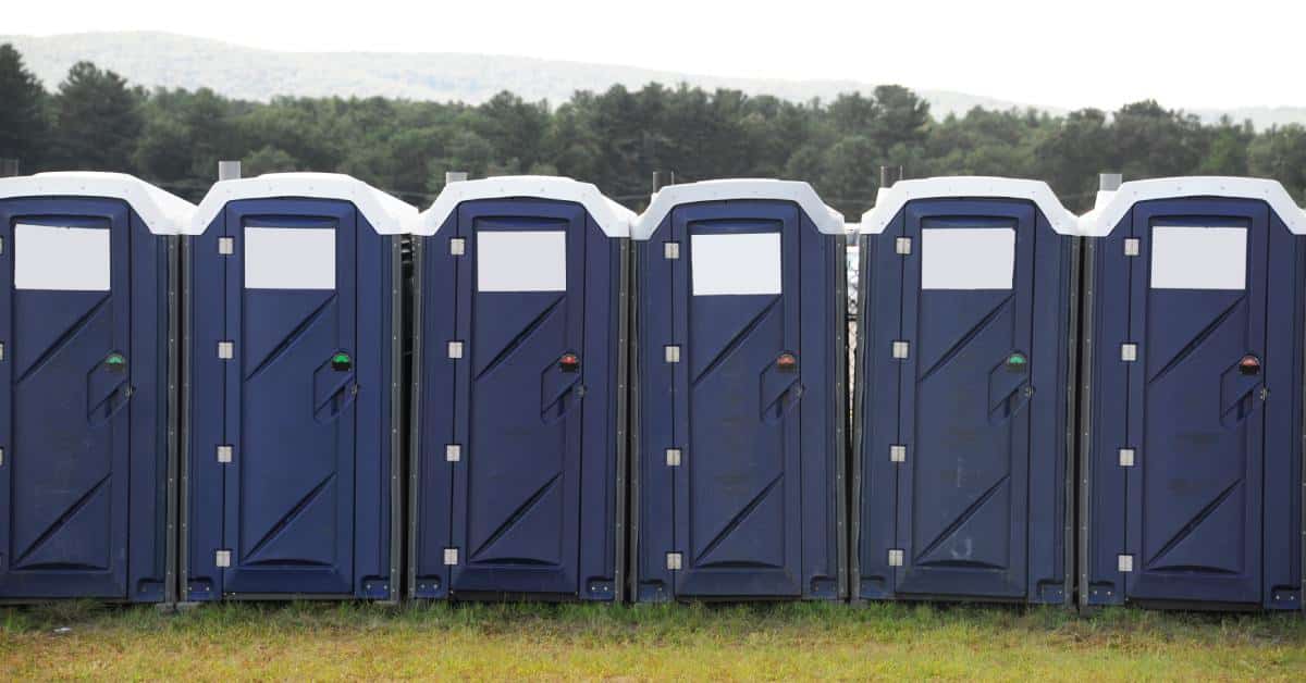 A row of six navy-blue portable toilets with white roofs positioned on a grassy field, with trees visible in the background.