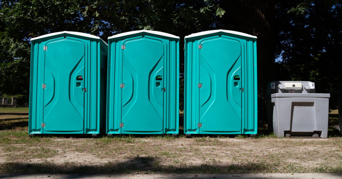 Three porta-potties lined up together in a sunny spot of grass. Next to the last one is a hand-washing station.
