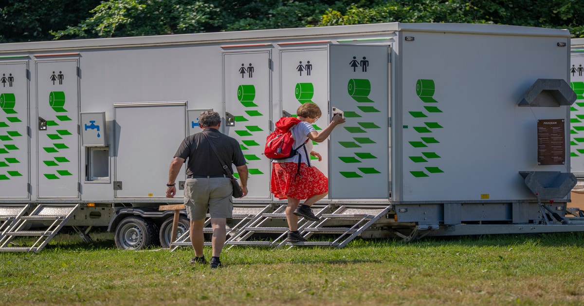 A man and a woman walking towards trailer bathrooms in a field of grass. The trailer features male and female symbols.