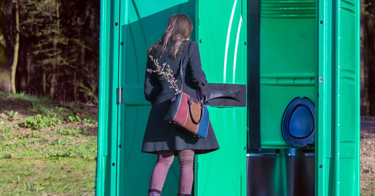 A woman wearing a purple dress and tights enters a green porta potty set on a patch of dirt near a wooded area.