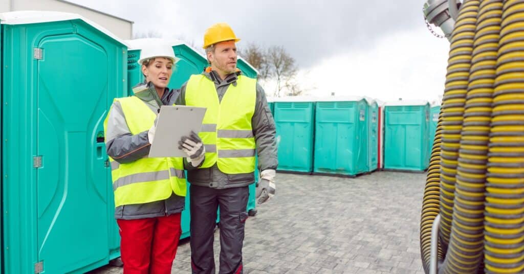 Two workers stand outside in front of rows of porta potties. One holds a clipboard, while the other looks at a coiled hose.
