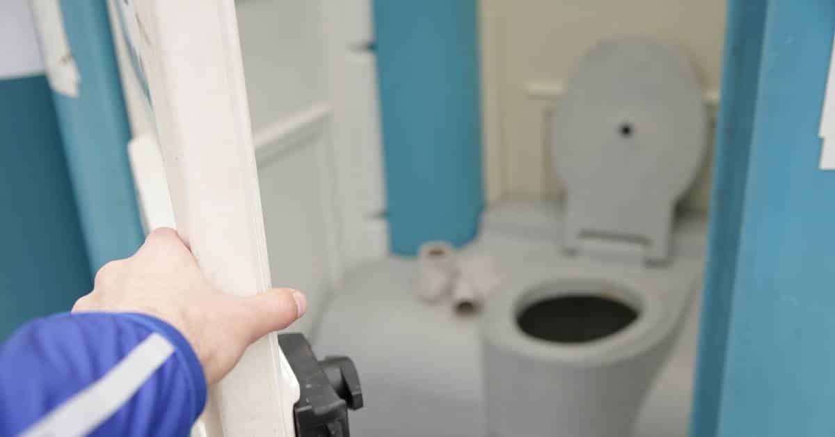 a white man's hand is reaching out to open the white and blue porta potty's door. The toilet is seen inside but out of focus.