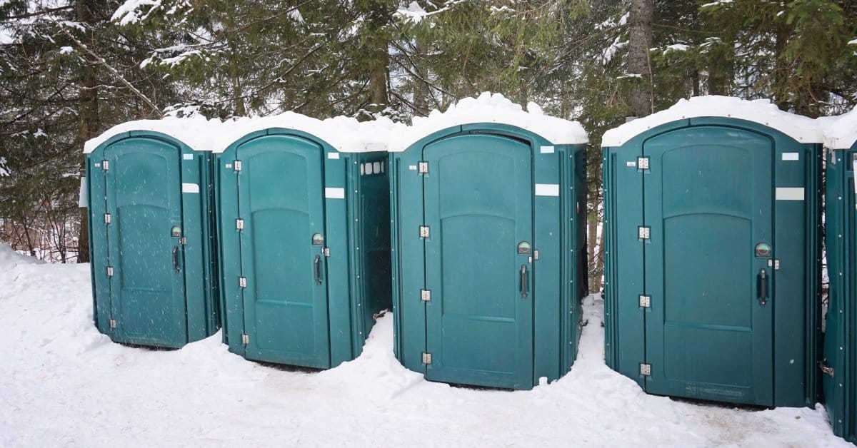 Five green Porta Potties with snow piled on top and lined up along the edge of a snow-covered forest of Pine trees.