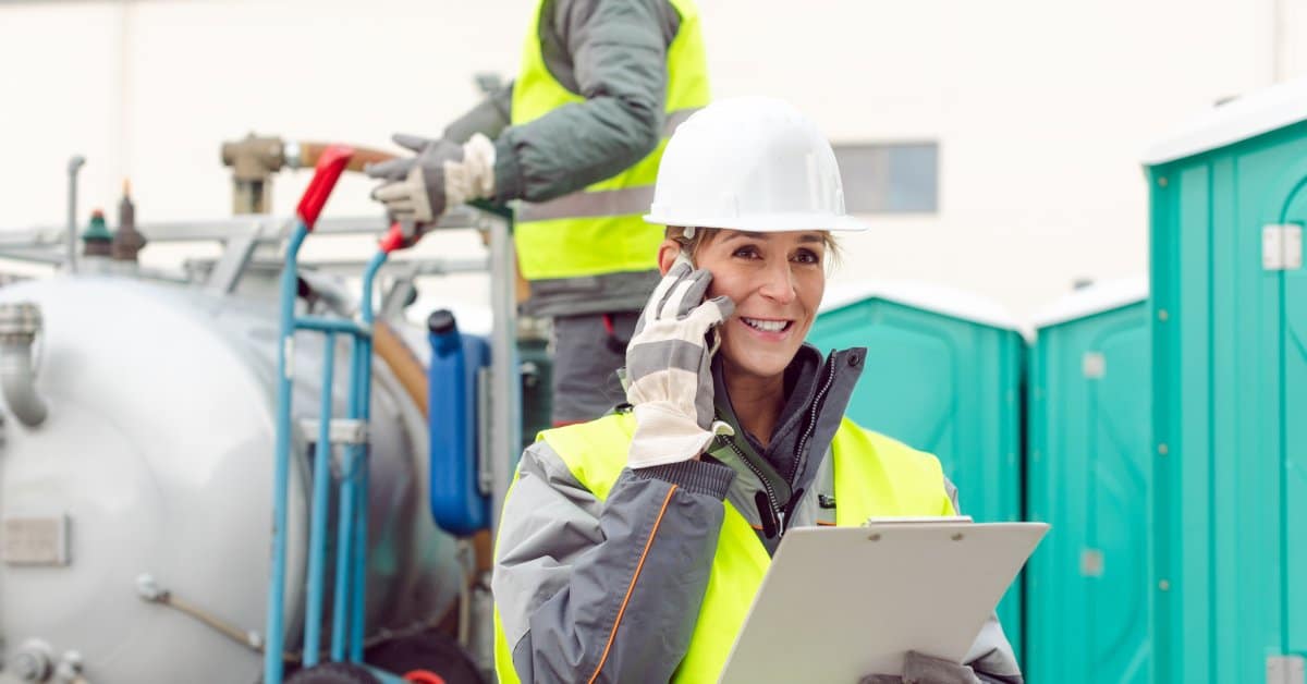 A woman and man in high-visibility vests and hard hats. The woman is on the phone and portable toilets are behind her.