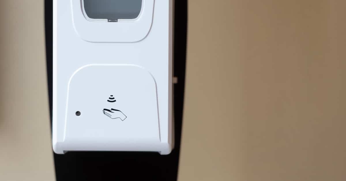 A close-up of a white automatic hand sanitizing station in front of a beige wall. The station has an icon of cupped hands.