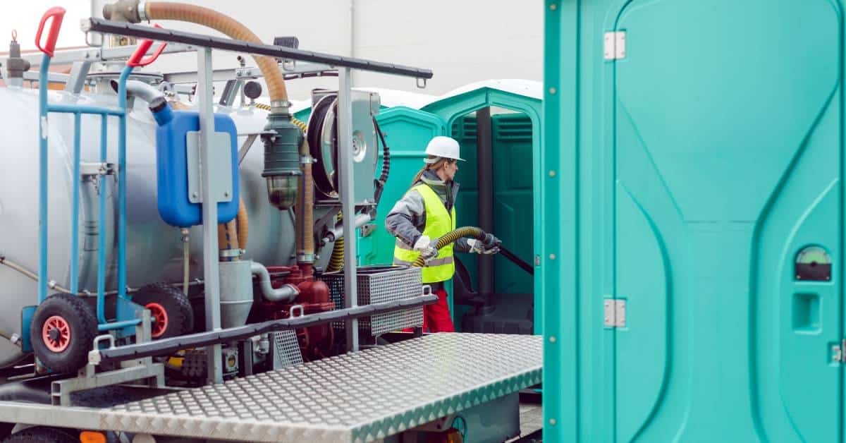 A woman in a high visibility vest holding a hose attached to a containment truck and cleaning out portable toilet waste.